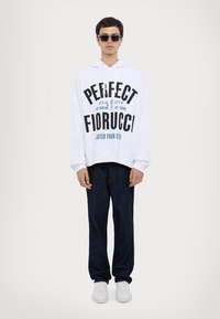 Hvid hoodie med sort og teal tekst, prydet med et grafisk print. Parret med mørke denimbukser og hvide sneakers, bærer modellen solbriller.