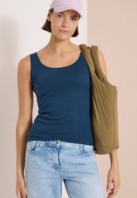 Mujer con una camiseta sin mangas azul marino, vaqueros azul claro y gorra rosa, lleva un bolso de hombro acolchado de color verde oliva.