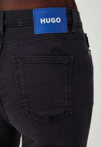 Svarte denimjeans med en blå lærmerkel med teksten "HUGO." Funksjoner inkluderer en baklomm og detaljer i sømmen.