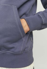 Felpa blu navy con finitura testurizzata, dotata di tasca frontale e polsini a costine. Sono visibili dettagli di cucitura notevoli.