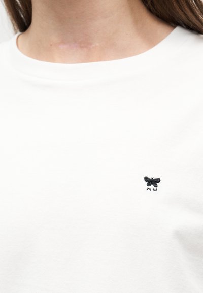 Κοντινό πλάνο ενός ατόμου που φοράει λευκό πουκάμισο με ένα μικρό μαύρο λογότυπο πεταλούδας στο στήθος και μια ελαφριά ουλή στον λαιμό πάνω από το κολάρο.