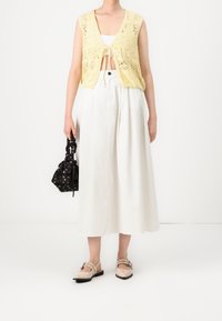 Keltainen neulottu liivi, valkoinen lyhyt toppi, pitkä valkoinen hame, musta kukkakuvioinen laukku ja luonnonväriset pukukengät solkikoristeella.