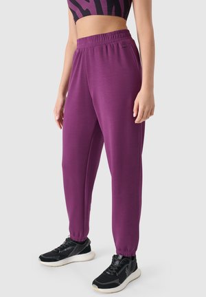 Personne portant un pantalon de jogging violet et des baskets noires avec des semelles blanches, debout devant un fond uni gris clair.