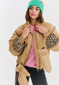 Mladá žena oblečená v béžovej pufferovej bunde s rukávmi s leopardím potlačou, ružovom svetre, mätovozelenom čiapke a pripevnených béžových rukaviciach, hľadí do strany.