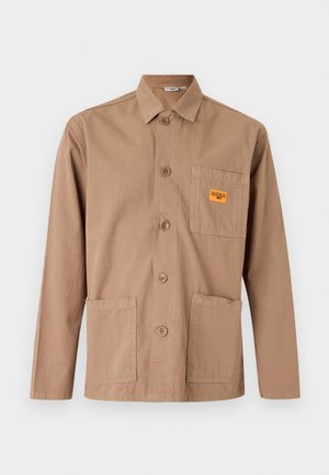 Pikkade varrukatega beež nööpidega jakk kraega, kolme esiosa taskuga ja väikese oranži plaastriga rindkere taskul.