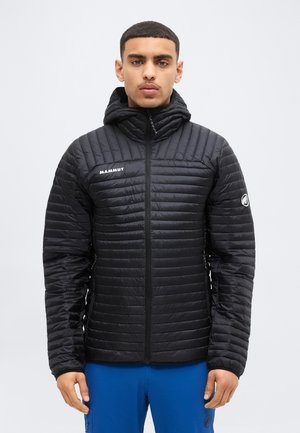 Νεαρός άνδρας που φοράει μαύρο καπιτονέ μπουφάν Mammut με κουκούλα και μπλε παντελόνι, στέκεται μπροστά σε απλό φωτεινό φόντο.