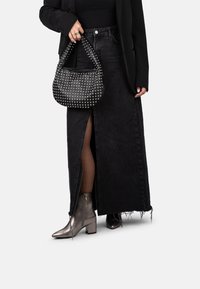 Una persona viste una camiseta sin mangas negra, una chaqueta ajustada y una falda de mezclilla larga, sostiene un bolso con tachuelas, combinado con botas metálicas hasta el tobillo.