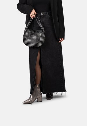 Una persona viste una camiseta sin mangas negra, una chaqueta ajustada y una falda de mezclilla larga, sostiene un bolso con tachuelas, combinado con botas metálicas hasta el tobillo.