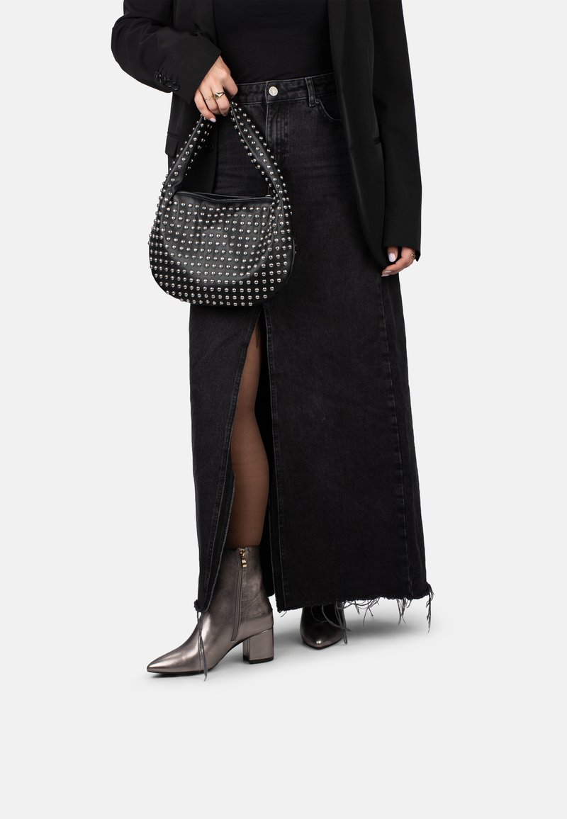 Una persona viste una camiseta sin mangas negra, una chaqueta ajustada y una falda de mezclilla larga, sostiene un bolso con tachuelas, combinado con botas metálicas hasta el tobillo.