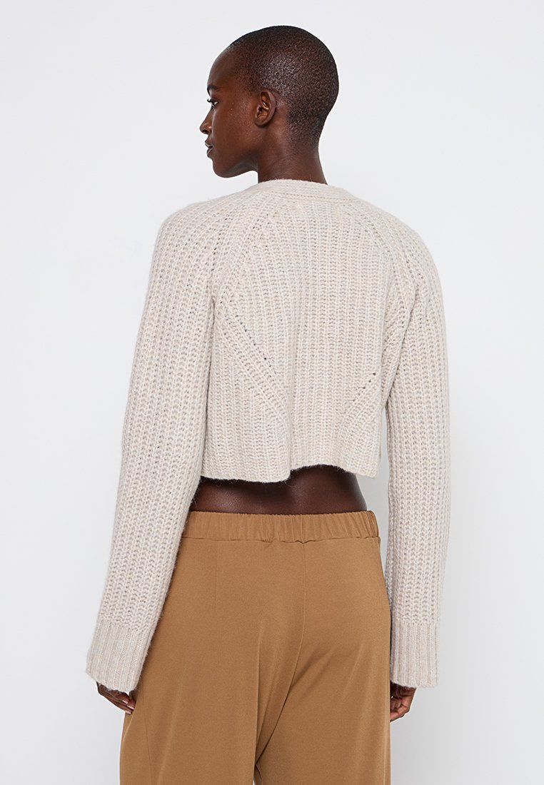 Beige gestrickter Pullover mit strukturiertem Muster und langen Ärmeln, getragen über braunen Hosen, der ein einfaches und gemütliches Design zeigt.