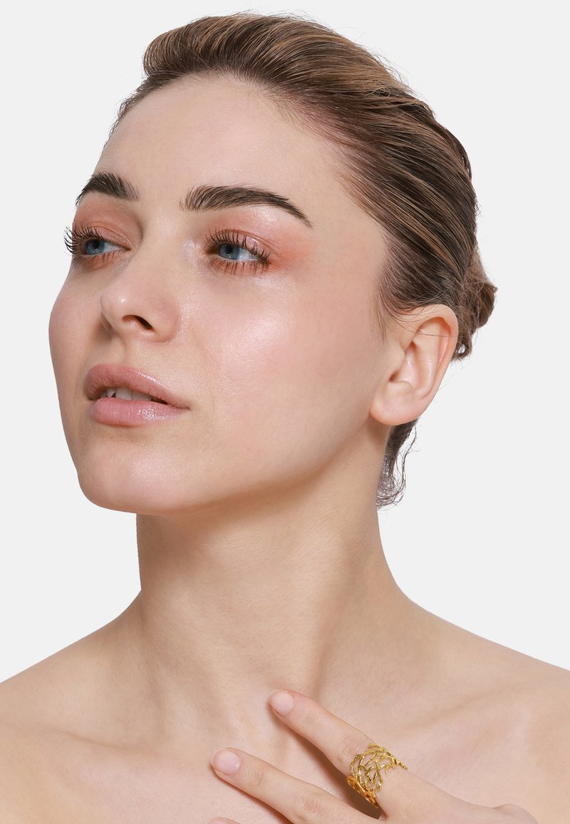 Gouden ring met een ingewikkeld geweven ontwerp, gedragen aan een vinger. Model heeft een egale huid, lichte make-up en een bun kapsel. Neutrale achtergrond.