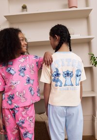 Zwei Kinder tragen passende Pyjama-Sets. Das erste Kind trägt ein pinkes Oberteil mit Stitch-Motiven und blaue Hosen; das zweite ein cremefarbenes Oberteil mit blauen Akzenten und hellblaue Hosen.
