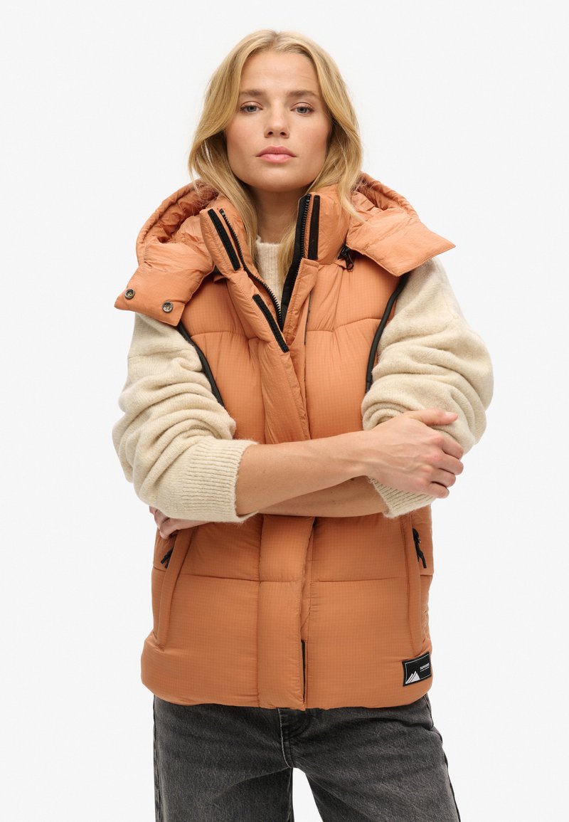 Pomarańczowa kamizelka puchowa z wysokim kołnierzem, zamkiem z przodu, dwoma bocznymi kieszeniami i czarnymi akcentami, nałożona na kremowy sweter.