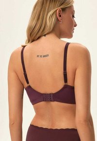 Biustonosz w ciemnym burgundzie, z regulowanymi ramiączkami, zapięciem na haczyk i oczy, gładkim materiałem oraz delikatnym tatuażem widocznym nad pasem.