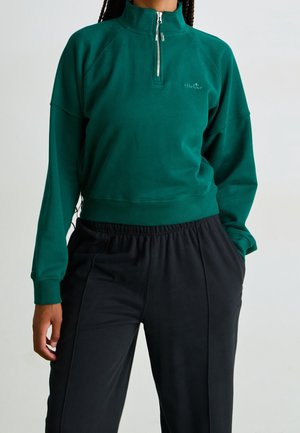 Person trägt ein grünes Viertel-Zip-Sweatshirt und schwarze Hose mit Händen in den Taschen, steht vor einem einfarbigen Hintergrund.