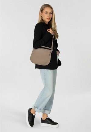 Vrouw draagt een zwarte blazer, lichtblauwe opgerolde spijkerbroek, zwarte sneakers, en draagt een beige schoudertas aan haar rechterschouder tegen een effen achtergrond.