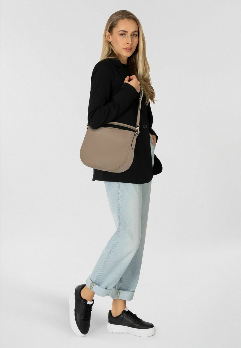 Femme portant un blazer noir, un jean bleu clair retroussé, des baskets noires, avec un sac à bandoulière beige sur l'épaule droite, devant un fond uni.