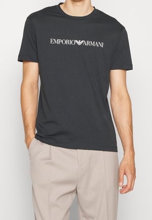 Man wearing a black Emporio Armani T-shirt and beige pants with one hand in pocket against a plain background.