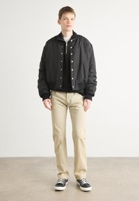 Jaqueta bomber preta com botões de pressão prateados, sobre um suéter preto. Calças bege claras e tênis pretos com solas brancas. Design simples.