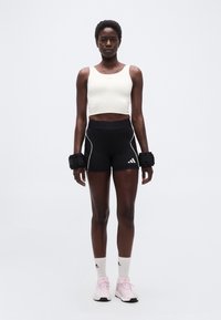 Αθλητική γυναίκα που φοράει λευκό τοπ κοντό, μαύρα σορτς, βάρη καρπού, λευκές κάλτσες και ροζ παπούτσια για τρέξιμο, στέκεται μπροστά σε απλό φόντο.