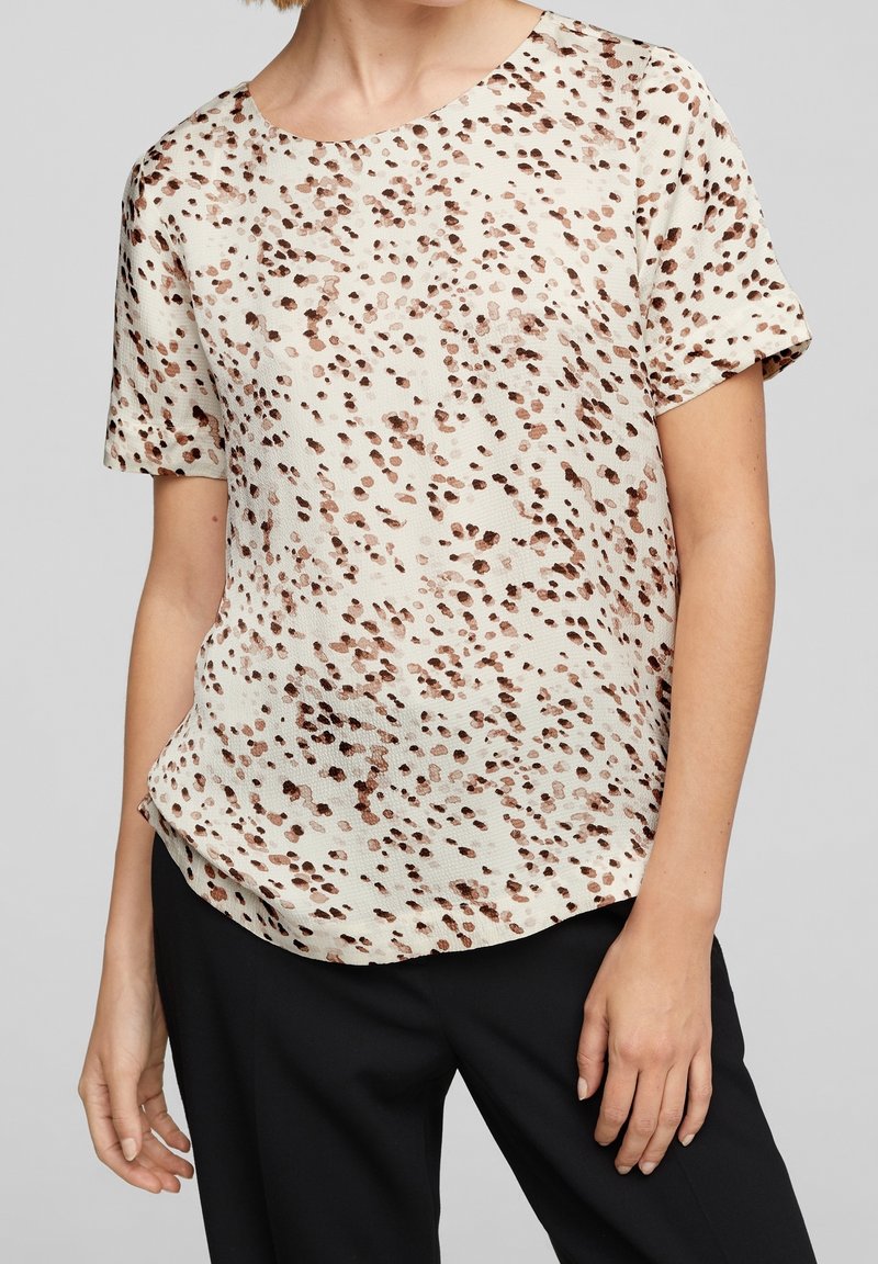 Femme portant un chemisier beige à manches courtes avec un motif de points irréguliers marron et un pantalon noir sur un fond gris uni.