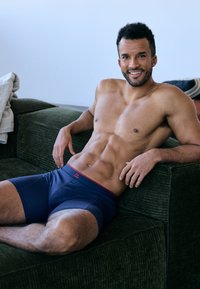 Námořnicky modré boxerky s červeným páskem, hladká textura a mírný lesk. Model sedí na zelené pohovce, ukazuje vypracovanou postavu.