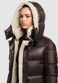 Brauner Puffermantel mit einem weichen, cremfarbenen Fleecefutter und übergroßer Kapuze. Verfügt über einen Reißverschluss vorne, Druckknöpfe und seitliche Taschen.