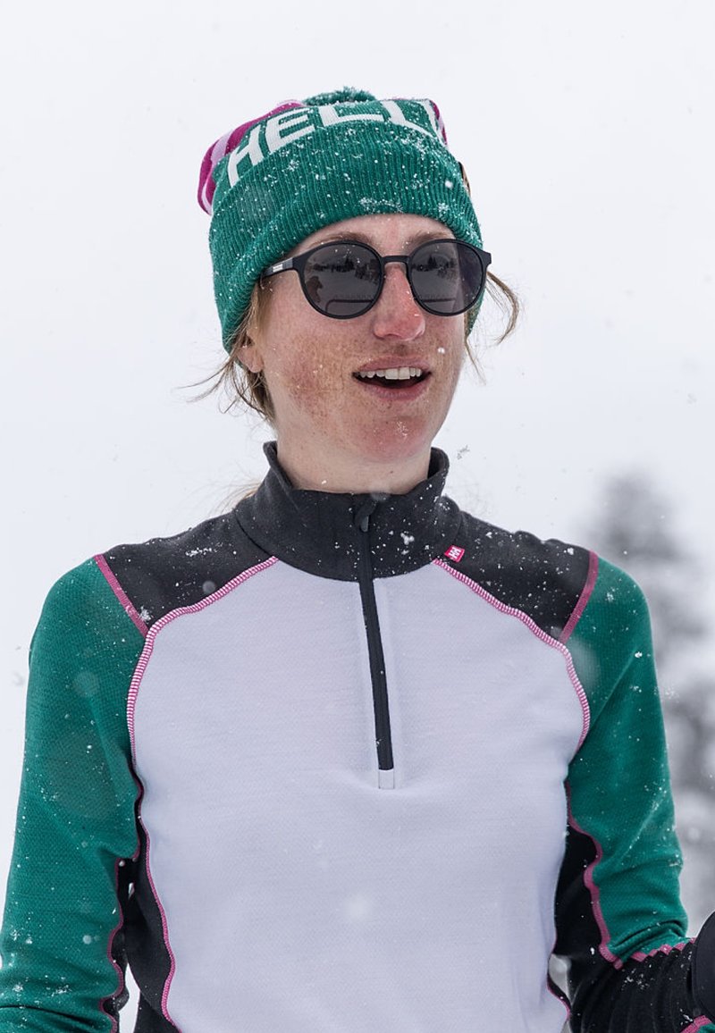 Gorro verde y rosa, gafas de sol redondas y una camiseta de manga larga con cuerpo blanco y mangas verdes, con costuras rosas, en la nieve.
