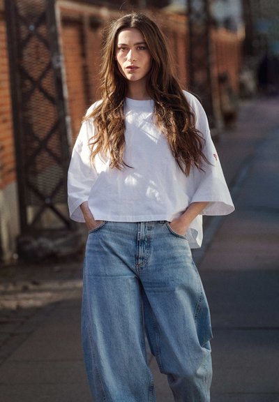 Bijela skraćena majica sa širokim rukavima u kombinaciji sa prostranim svijetloplavim trapericama. Tkanina izgleda mekano, s vidljivim detaljima šavova.