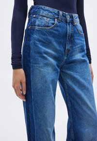 Τζιν ψηλόμεσα παντελόνια σε μεσαίο έως σκούρο μπλε χρώμα, με κάθετα πλαϊνά πάνελ σε αντίθεση και πέντε τσέπες με χαλκινα κουμπιά.