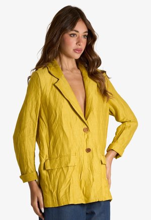 Donna con lunghi capelli castani che indossa un blazer giallo, ampio e strutturato, con due bottoni e pantaloni blu, posando con una mano sul fianco.