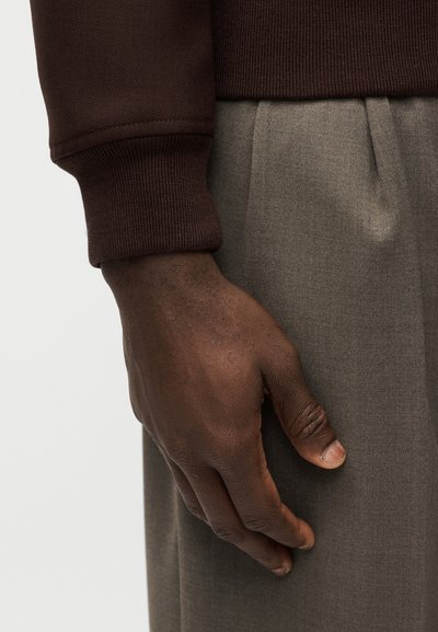 Χέρι με σκουρόχρωμο δέρμα που ξεκουράζεται κοντά στην τσέπη γκρι πλισέ παντελονιού, φορώντας καφέ πουλόβερ με μακριά μανίκια και ριμπ τελείωμα.