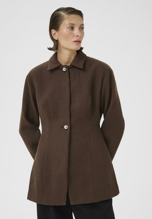 Femme aux cheveux courts portant une veste marron côtelée avec des boutons argentés, debout les mains dans le dos, regardant vers la droite sur un fond uni.