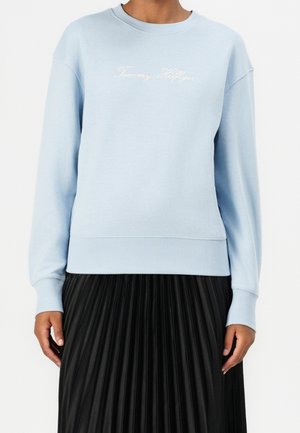Lyseblå sweatshirt med teksten "Tommy Hilfiger" båret med sort plisseret nederdel, vist fra hals til hofter mod ensfarvet baggrund.