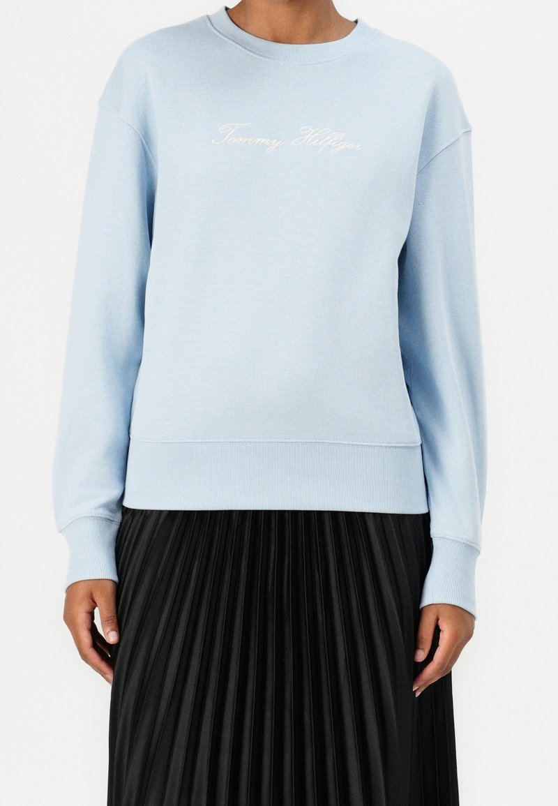 Sweat-shirt bleu clair avec le texte « Tommy Hilfiger » porté avec une jupe plissée noire, visible du cou aux hanches sur un fond uni.