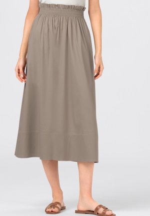 Femme portant une jupe midi taille haute marron clair avec une ceinture à volants et des sandales plates marron, debout devant un fond uni.