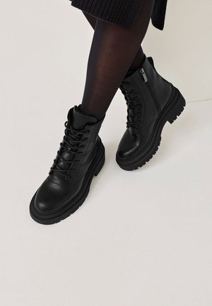 Czarne skórzane wojskowe buty na sznurowadła noszone z czarnymi rajstopami i czarną spódnicą na jasnym tle.