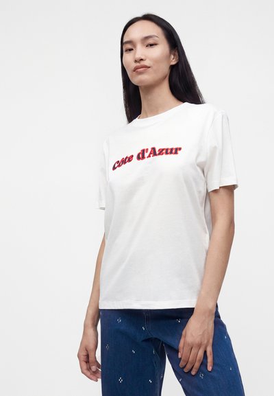 Femme aux longs cheveux foncés portant un t-shirt blanc avec un texte rouge « Cote d'Azur » et un jean bleu avec de petits motifs blancs, debout devant un fond blanc.