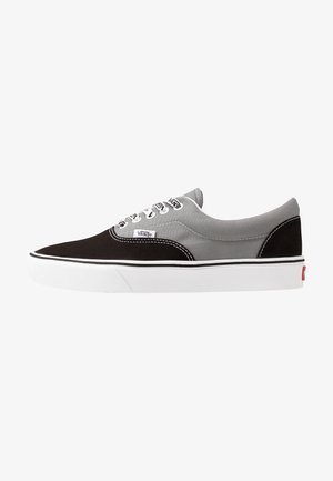 Baskets basses Vans en toile noire et grise avec lacets blancs, semelle en caoutchouc blanche, et étiquettes logo Vans sur le côté et la languette.