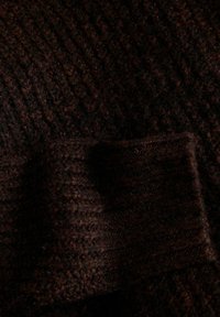 Gros plan sur un tissu tricoté marron foncé avec une surface texturée et duveteuse et des motifs ainsi que des points visibles.