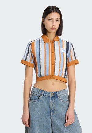 Vrouw draagt een kort, gebreid T-shirt met korte mouwen en verticale strepen in oranje, blauw, wit en zwart, gecombineerd met losse blauwe spijkerbroeken.