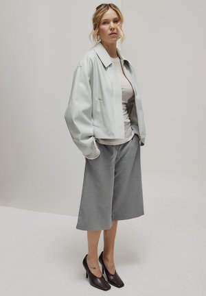Femme debout portant une veste ample gris clair, un haut beige, des culottes larges grises et des talons hauts marron foncé sur fond uni.