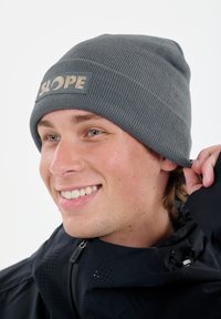Grijze gebreide beanie met een geribbelde textuur en een rechthoekige patch met het woord "SLOPE" in beige. De persoon draagt een donkere jas.