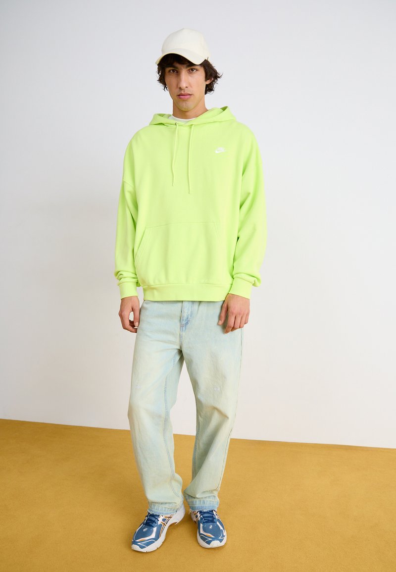 Felpa verde neon con cappuccio e coulisse, tasca frontale e logo bianco; abbinata a jeans in denim azzurro chiaro e sneaker blu/bianco, modello con cappello.
