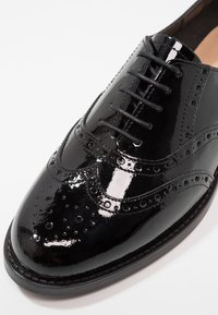Chaussure oxford à bouts fleuris en cuir verni noir avec perforations de brogue et fermeture à lacets sur un fond blanc.