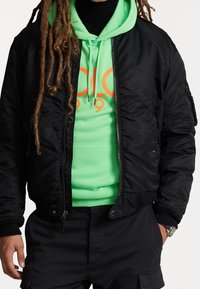 Svart bomberjacka med ribbade ärmar, över en klargrön hoodie med en orange grafisk design. Strukturerad tyg och dragkedja synliga.