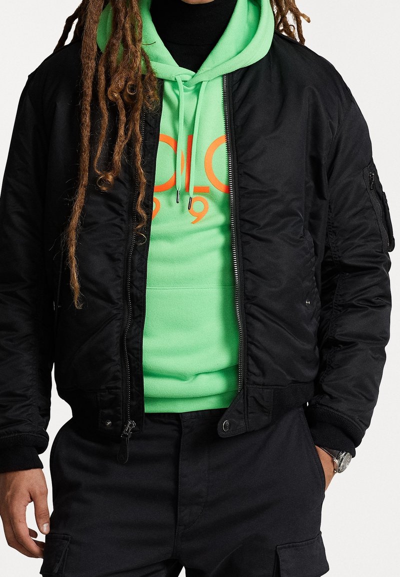 Svart bomberjacka med ribbade ärmar, över en klargrön hoodie med en orange grafisk design. Strukturerad tyg och dragkedja synliga.