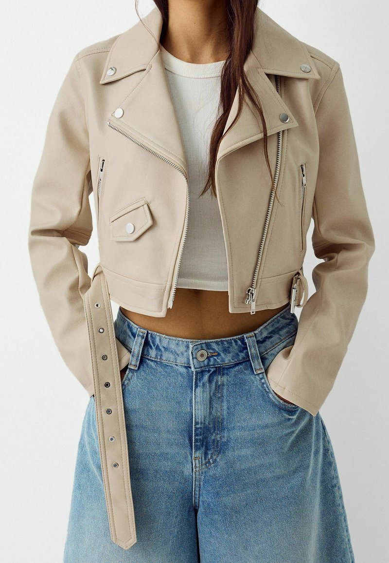 Giacca da motociclista corta in ecopelle beige con chiusura a zip, tasche laterali e bottoni a pressione; abbinata a short di jeans blu a vita alta.