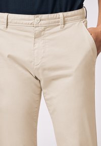 Beige chinos made of lightweight fabric featuring a flat front, side pockets, and a classic button closure, with subtle stitch detailing.