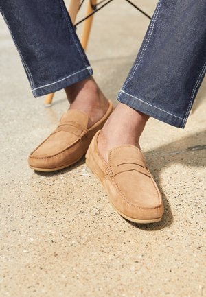 Mocassins en daim de couleur tan avec un design de mocassins, présentant des accents cousus et une texture douce. Associés à un pantalon en denim foncé.